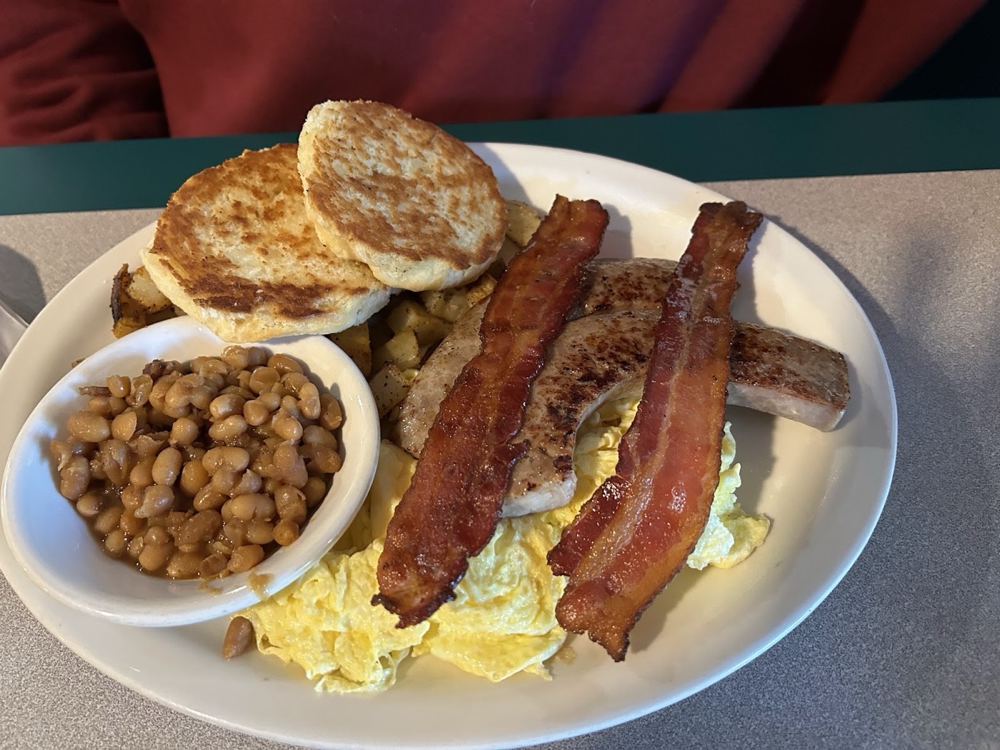 A diner breakfast of scrambled eggs, bacon, sausage, home fries, baked beans and a grilled English muffin.